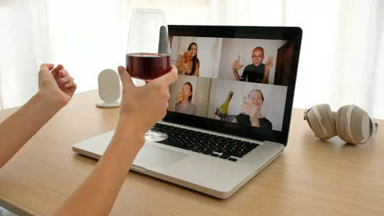 Woman drinks wine in front of laptop showing video call.